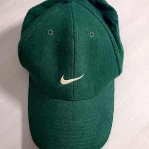 May include: A dark green baseball cap with a felt-like texture, featuring a white embroidered Nike swoosh logo on the front center. The cap includes a curved brim, a button on the top crown, and small ventilation eyelets stitched in matching green.