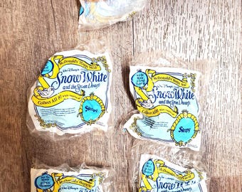 Può includere: Cinque bustine di plastica McDonald's Happy Meal con il marchio di Biancaneve e i sette nani. Una bustina contiene una statuetta di Cucciolo con tunica viola e barba bianca. Le altre quattro bustine sono sigillate con testo stampato in blu e giallo.