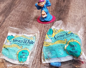 Puede incluir: Dos bolsas de plástico que contienen pequeños juguetes de plástico de una promoción de Happy Meal de McDonald's para la película Blancanieves y los Siete Enanitos. Las bolsas están etiquetadas como "Blancanieves y los Siete Enanitos" y presentan a los personajes Dopey y Sneezy. Los juguetes son azules y rojos.