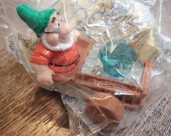 May include: A plastic figurine of the character Doc from Snow White and the Seven Dwarfs, wearing a green hat and orange shirt, pushing a brown cart filled with clear and teal-colored plastic gems, all sealed inside a clear plastic bag.