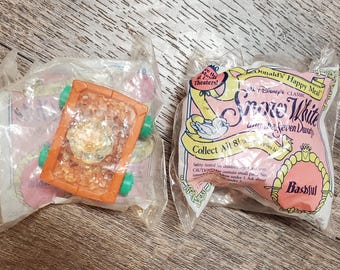 May include: A plastic bag containing a small toy carriage with a brown and orange body and green wheels. The bag also contains a pink and white card with the text "Donald's Happy Meal", "Disney's Classic", "Snow White and the Seven Dwarfs", "Collect All 8", and "Bashful".