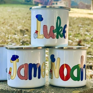 May include: White enamel mugs with silver rims, each personalized with a name in colorful, playful lettering. One mug is blank. The names include "Luke", "James", and "Noah", with decorative accents.