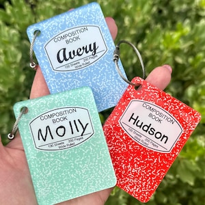 May include: Three composition book-style keychains in blue, green, and red. Each keychain features a white label with a name and the words "Composition Book", "100 Sheets", "200 Pages", and "Wide Ruled". The names are Avery, Molly, and Hudson.