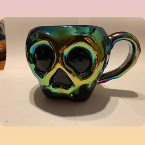 May include: A black iridescent ceramic mug shaped like a skull with a handle. The mug has a glossy finish and is decorated with a skull design.