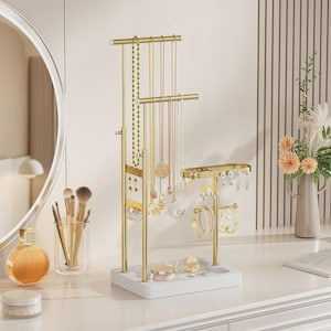 May include: A gold-toned metal jewelry organizer stand with a white rectangular base. The stand features multiple tiers for hanging necklaces, a perforated panel for earrings, and a horizontal bar for bracelets, holding various pieces of jewelry.