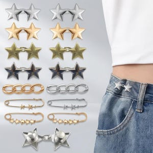 May include: A collection of metal clothing fasteners including double star pins in silver, gold, and bronze tones, chain-link pins, and safety pins adorned with small star charms. One silver double star pin is shown attached to blue denim jeans.