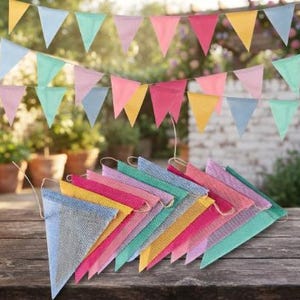 May include: A decorative bunting banner featuring triangular flags in pastel shades of pink, yellow, blue, and green. The set includes a string of hanging flags and a stacked pile of matching triangular pennants with a textured, woven appearance.