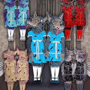 May include: Enamel pins featuring a wolf-like creature in a dress, inspired by a horror film. The pins are in various colors, including blue, red, gold, and black. The wolf has a menacing expression with sharp teeth and a lolling tongue. The pins are arranged in pairs.