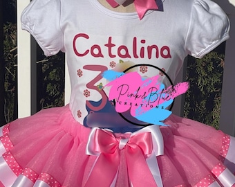 May include: A white short-sleeved top with the name "Catalina" and the number "3" in red, combined with a pink tutu skirt. The skirt features white and pink polka dot ribbons and a large bow. A matching bow headband completes the ensemble.