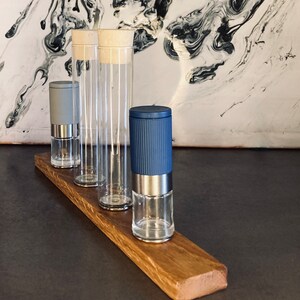 Könnte beinhalten: Vier gläserne Gewürzmühlen auf einem Holzständer. Zwei Mühlen sind aus klarem Glas mit einem Holzdeckel. Eine Mühle ist blau mit einem silbernen Deckel. Eine Mühle ist grau mit einem silbernen Deckel.