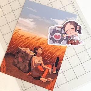 May include: A rectangular art print and a die-cut sticker featuring a character with brown hair in a white tank top and orange pants sitting in a wheat field next to a grey cube with a pink heart. The art print shows the character leaning against the cube.