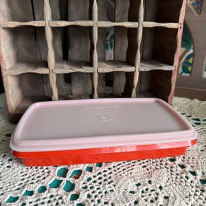 Op de afbeelding: Een rechthoekige Tupperware-opbergdoos met een felrode basis en een doorschijnend, lichtroze deksel. Het deksel is voorzien van een gecentreerd rond embleem en de in reliëf aangebrachte merknaam Tupperware. Plat en laag ontwerp.