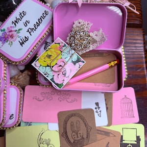 May include: A collection of stationery items, including pink tins with floral and rhinestone accents, decorative paperclips, and small cards with illustrations. The text "Write in His Presence" is visible on one tin.