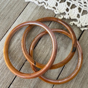 Puede incluir: Dos brazaletes circulares con un acabado liso y pulido. Las pulseras presentan un color ámbar marrón cálido y translúcido con sutiles vetas jaspeadas de color marrón oscuro en todo el material, creando un aspecto estratificado y semitransparente.