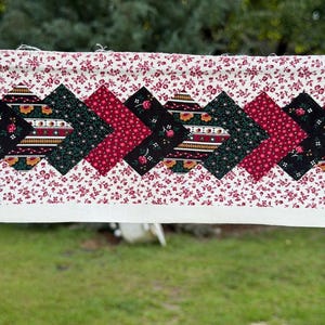 May include: A rectangular fabric strip featuring a central row of interconnected diamond-shaped patches in black, dark green, and deep red with floral and striped patterns. The strip is bordered by a cream-coloured fabric with a small red floral print.