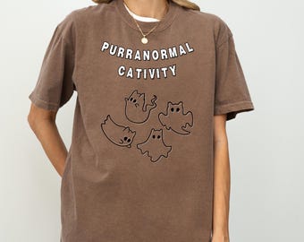 May include: Brown t-shirt with the text "PURRANORMAL CATIVITY" above four cartoon cat ghost illustrations. The shirt has a crew neck and short sleeves.