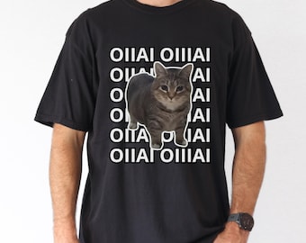 Può includere: T-shirt nera con grafica di gatto tigrato. Il gatto è al centro, con il testo "OIIAI" impilato verticalmente su entrambi i lati. La maglietta è a girocollo.