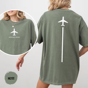May include: Moss green t-shirt with a white airplane graphic and the text "AIRPLANE MODE" on the front. The back of the shirt features a white airplane graphic with a white line extending downwards. The shirt is a relaxed fit.
