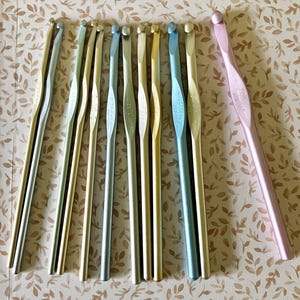 May include: A collection of crochet hooks in various metallic colors, including silver, gold, and blue, with a single pink hook. The hooks are arranged on a patterned surface, showcasing their different sizes and styles. The hooks are made in the USA.