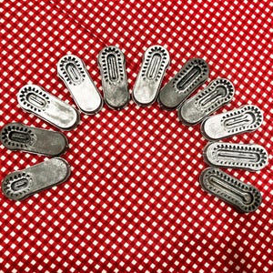May include: A collection of silver-toned metal shoe-shaped charms arranged in a semi-circle on a red and white checkered fabric. Each charm has a detailed sole design.