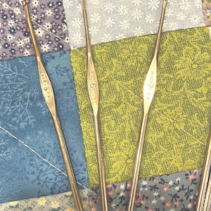 May include: Close-up of several vintage crochet hooks. The hooks are made of a shiny, silver-colored metal. The hooks are labeled with sizes and "MADE U.S.A." The background is a patchwork quilt with floral and geometric patterns.
