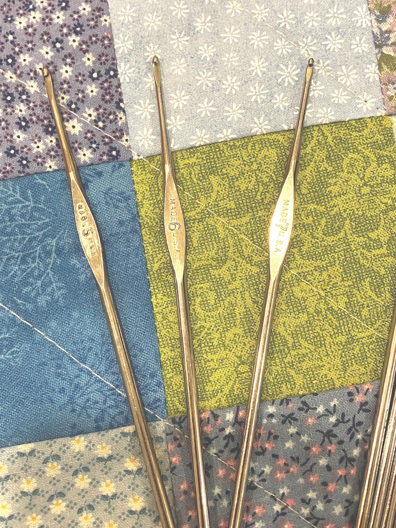 May include: Close-up of several vintage crochet hooks. The hooks are made of a shiny, silver-colored metal. The hooks are labeled with sizes and "MADE U.S.A." The background is a patchwork quilt with floral and geometric patterns.