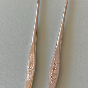 May include: Two silver crochet hooks. One hook is marked "MADE IN U.S.A.", and the other is marked "U.S.A. 10 1/2". The hooks are slender with a hook at the end for catching yarn. The background is a light blue-gray.