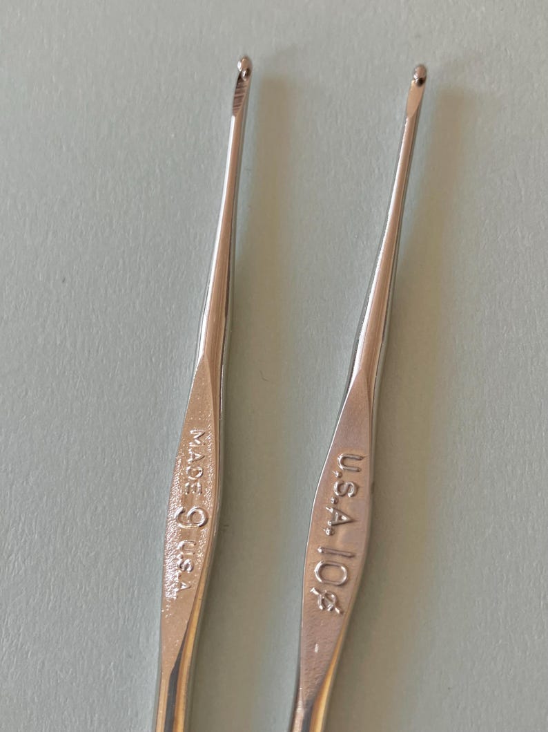May include: Two silver crochet hooks. One hook is marked "MADE IN U.S.A.", and the other is marked "U.S.A. 10 1/2". The hooks are slender with a hook at the end for catching yarn. The background is a light blue-gray.