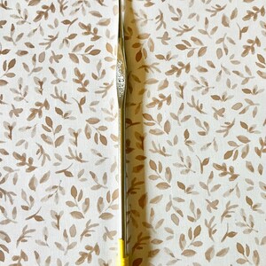May include: A silver crochet hook with a yellow handle. The hook is slender and has a pointed tip. The handle is a bright yellow color. The background is a cream-colored fabric with a repeating pattern of brown leaves.