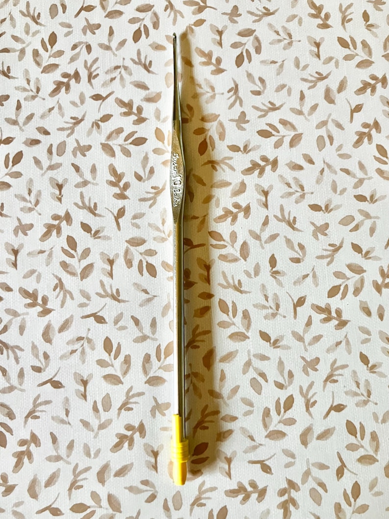 May include: A silver crochet hook with a yellow handle. The hook is slender and has a pointed tip. The handle is a bright yellow color. The background is a cream-colored fabric with a repeating pattern of brown leaves.