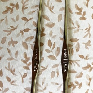 May include: Two silver crochet hooks with curved tips are displayed against a patterned background. One hook is marked "Susan Bates" and the other is marked "MADE | USA."