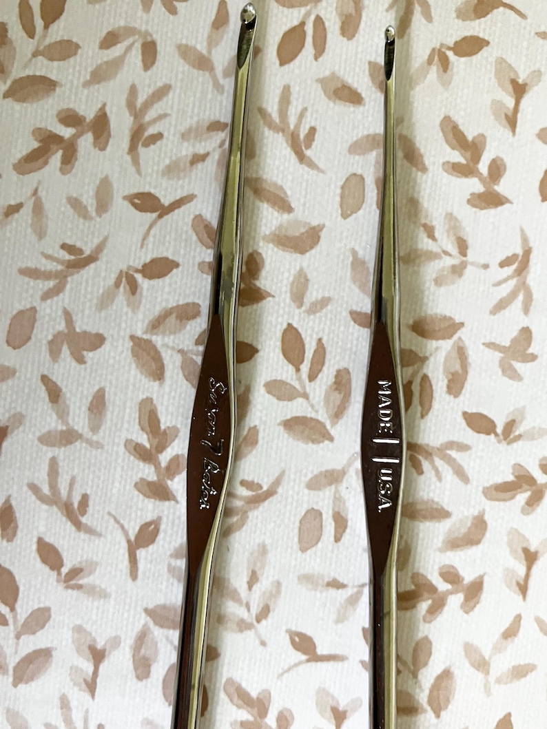 May include: Two silver crochet hooks with curved tips are displayed against a patterned background. One hook is marked "Susan Bates" and the other is marked "MADE | USA."