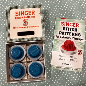 May include: A Singer Stitch Patterns for Automatic Zigzagger set including a square box with four blue plastic cams and a printed instruction booklet. The box lid and booklet feature the Singer logo and text detailing the stitch patterns.