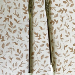 May include: Two crochet hooks with silver metal shafts and white plastic handles. The hooks are positioned vertically against a background of a beige and brown floral pattern. One hook has the text "Susan Bates" and the other has "MADE | USA".