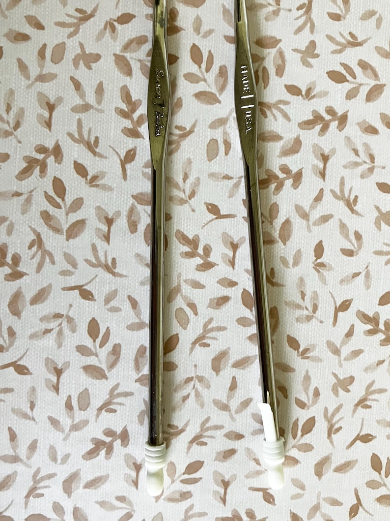May include: Two crochet hooks with silver metal shafts and white plastic handles. The hooks are positioned vertically against a background of a beige and brown floral pattern. One hook has the text "Susan Bates" and the other has "MADE | USA".