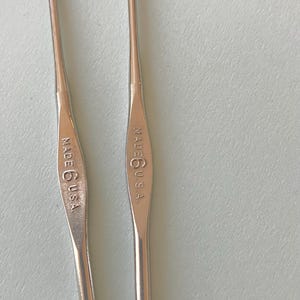 May include: Two silver crochet hooks. Each hook has a curved end and a handle with the text "MADE 6 U.S.A." stamped on it. The hooks are designed for yarn crafts and are set against a light blue background.
