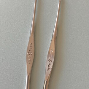 May include: Two silver crochet hooks. One hook is marked "MADE U.S.A." and the other is marked "Susan Bates". The hooks are slender with a curved tip for catching yarn. The background is a light, solid color.