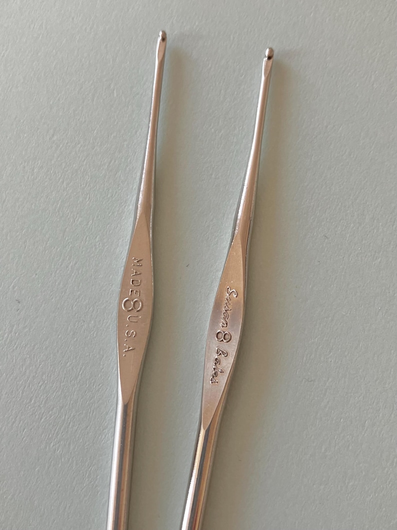May include: Two silver crochet hooks. One hook is marked "MADE U.S.A." and the other is marked "Susan Bates". The hooks are slender with a curved tip for catching yarn. The background is a light, solid color.