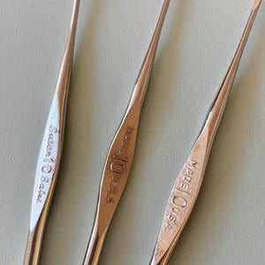 May include: Three silver crochet hooks. One hook has the text "Susan Bates" engraved on it. The other two hooks have the text "Made in USA" and the number "10" engraved on them. The hooks are made of metal.