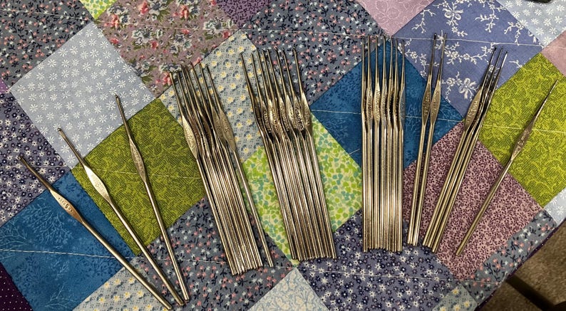 May include: A collection of silver crochet hooks of various sizes, arranged on a colorful patchwork quilt. The hooks are made of metal and have a slender, pointed design, ideal for yarn crafts. The quilt features a mix of floral and geometric patterns.