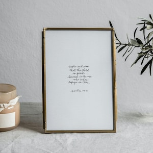 May include: A framed print with handwritten text, a lit candle in a glass jar with a wooden lid, and a small vase with greenery on a white surface. The print has a gold frame and the candle has a white ribbon.