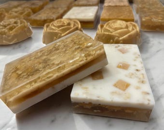 Puede incluir: Jabones artesanales en varias formas, incluyendo barras rectangulares y jabones con forma de rosa. Los jabones tienen un diseño en capas con una capa superior transparente, y algunos contienen ingredientes visibles. La paleta de colores es principalmente beige y blanco.
