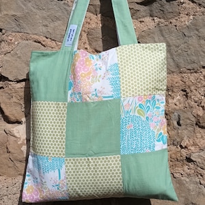 Peut inclure: Un sac cabas en patchwork vert avec des motifs floraux et géométriques. Le sac est muni d'un cintre en bois et d'une étiquette indiquant "Fabriqué avec amour".