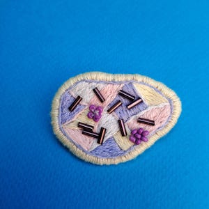 Peut inclure: Écusson brodé aux couleurs pastel, avec un motif géométrique composé de sections roses, lavande et jaunes. L'écusson est orné de petites perles marron foncé et de perles violettes en forme de fleur, le tout sur une bordure jaune clair.