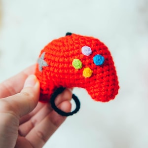 Può includere: Un controller per videogiochi rosso all'uncinetto fatto a mano con bottoni colorati e un anello nero. Il controller è tenuto in una mano, con un braccialetto visibile. Sullo sfondo ci sono gomitoli di lana rosa e blu.