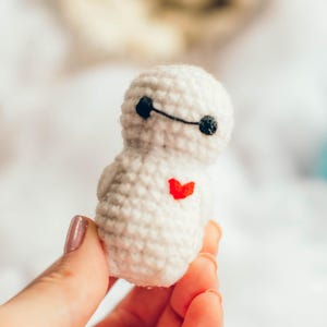 May include: A small, handmade white crochet figure with black eyes and a black line for a mouth, and a red heart on its chest. The figure is held in a hand against a soft, blurred background of white and colorful yarn.