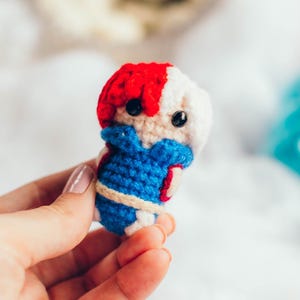 May include: A small, handmade crochet doll in the style of a character with red and white hair, blue and white outfit, and black eyes. The doll is held in a hand against a background of white fluff and colorful yarn.