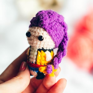 May include: A small, handmade crochet doll with a purple hat and long braid. The doll has black eyes, a beige face, and a yellow and blue outfit. The doll is held in a hand against a blurred background with colorful pom-poms.