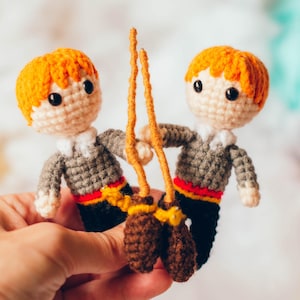 Puede incluir: Dos muñecos de ganchillo hechos a mano con pelo naranja, suéteres grises y pantalones negros. Sostienen una escoba marrón. Los muñecos tienen ojos negros y están hechos con hilo. El fondo incluye ovillos de lana en rojo y turquesa.