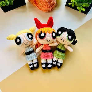 Puede incluir: Tres muñecas de crochet, cada una con un vestido y cabello de diferente color. Las muñecas sonríen y tienen ojos grandes y negros. Las muñecas están sentadas sobre una superficie marrón claro.